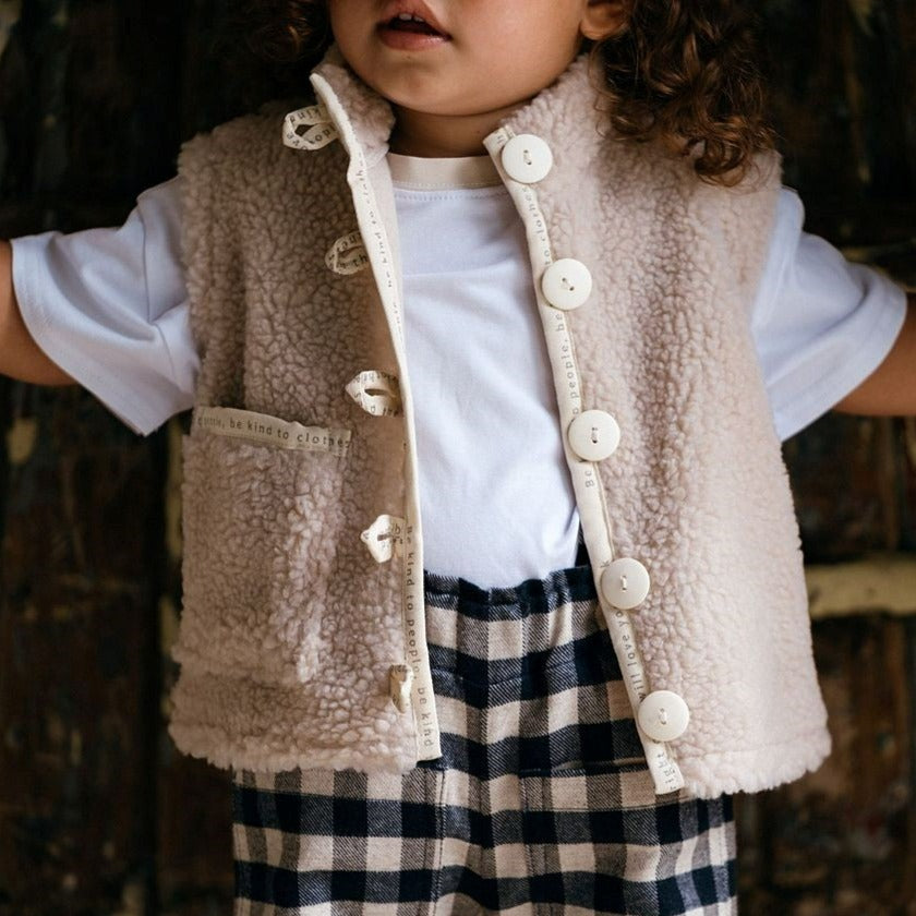 Brushed blue gingham cotton and wool unisex kids adjustable elasticated waisted trousers with an offwhite organic jersey short sleeve kids tee shirt with sand contrast collar.  Worn with a kids unisex beige faux fur gilet with contrast offwhite Lacuna Child engraved buttons and binding.