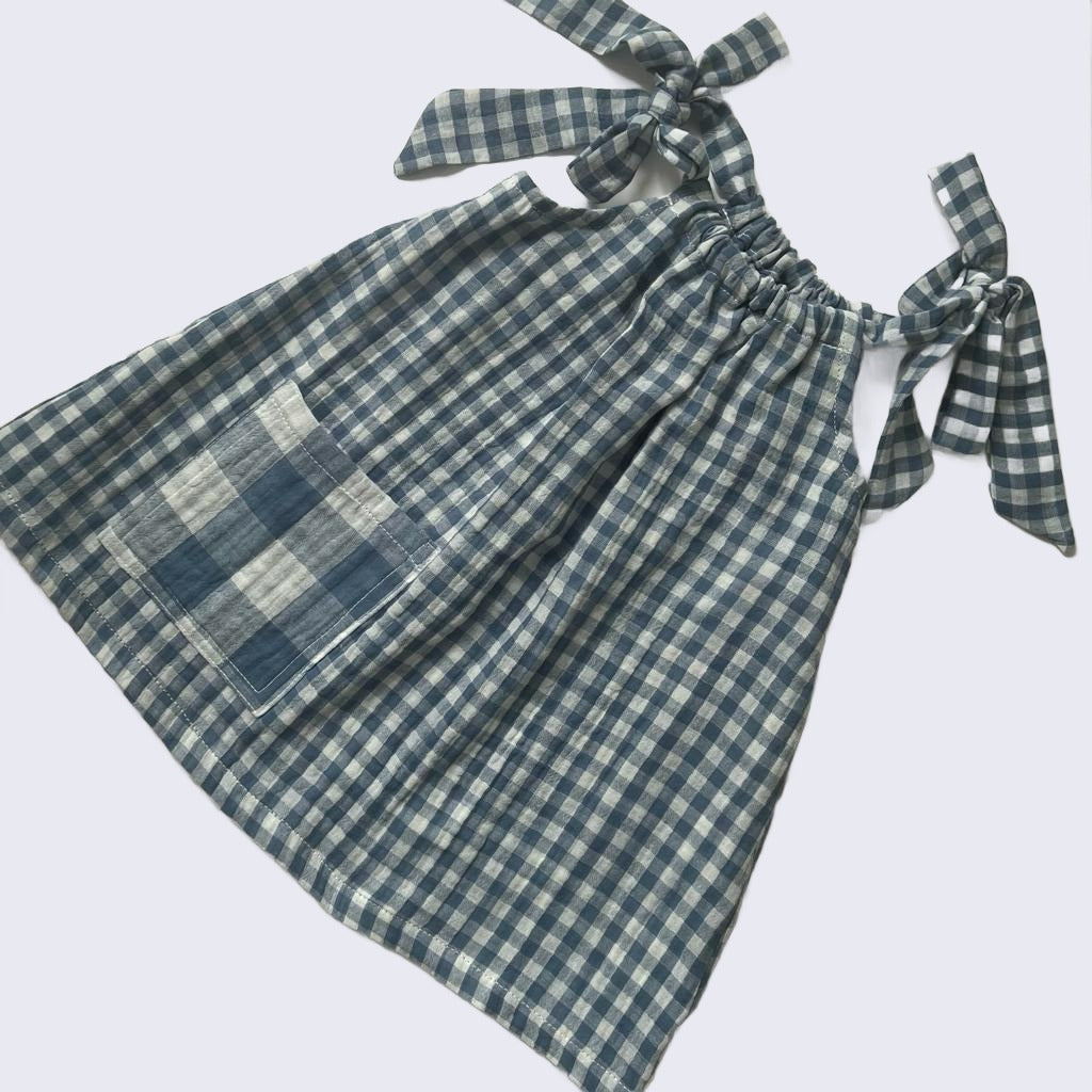 Flatlay on white background showing girls blue gingham dress with oversized pocket and bow tie straps