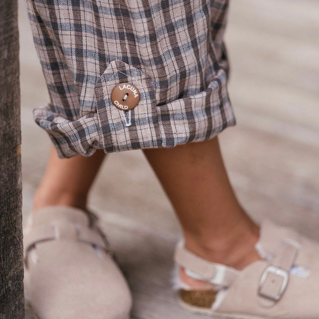 Childrens brown check linen trousers with tab to hold rolled up hem allowing for growing room.  Brown button holding tab engraved with the name Lacuna Child