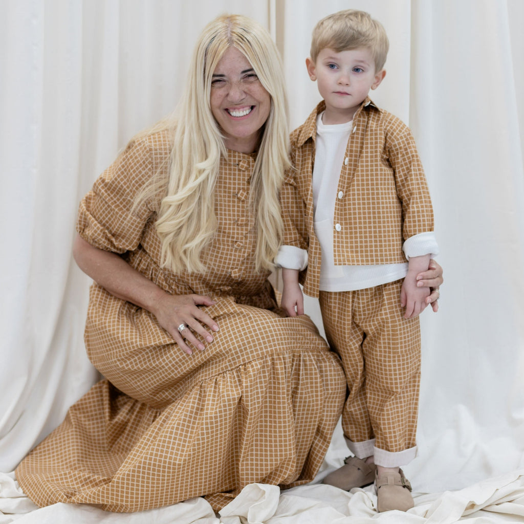 Ladies Betsy dress in caramel cotton with a scalloped front with covered button opening.  Crouched next to a boy wearing the Bailey cotton shirt in caramel with a white square print.  White jersey Buddy waffle top worn under with sleeves rolled over shirt.  Worn with Teddy caramel cotton square print jeans with hems turned up