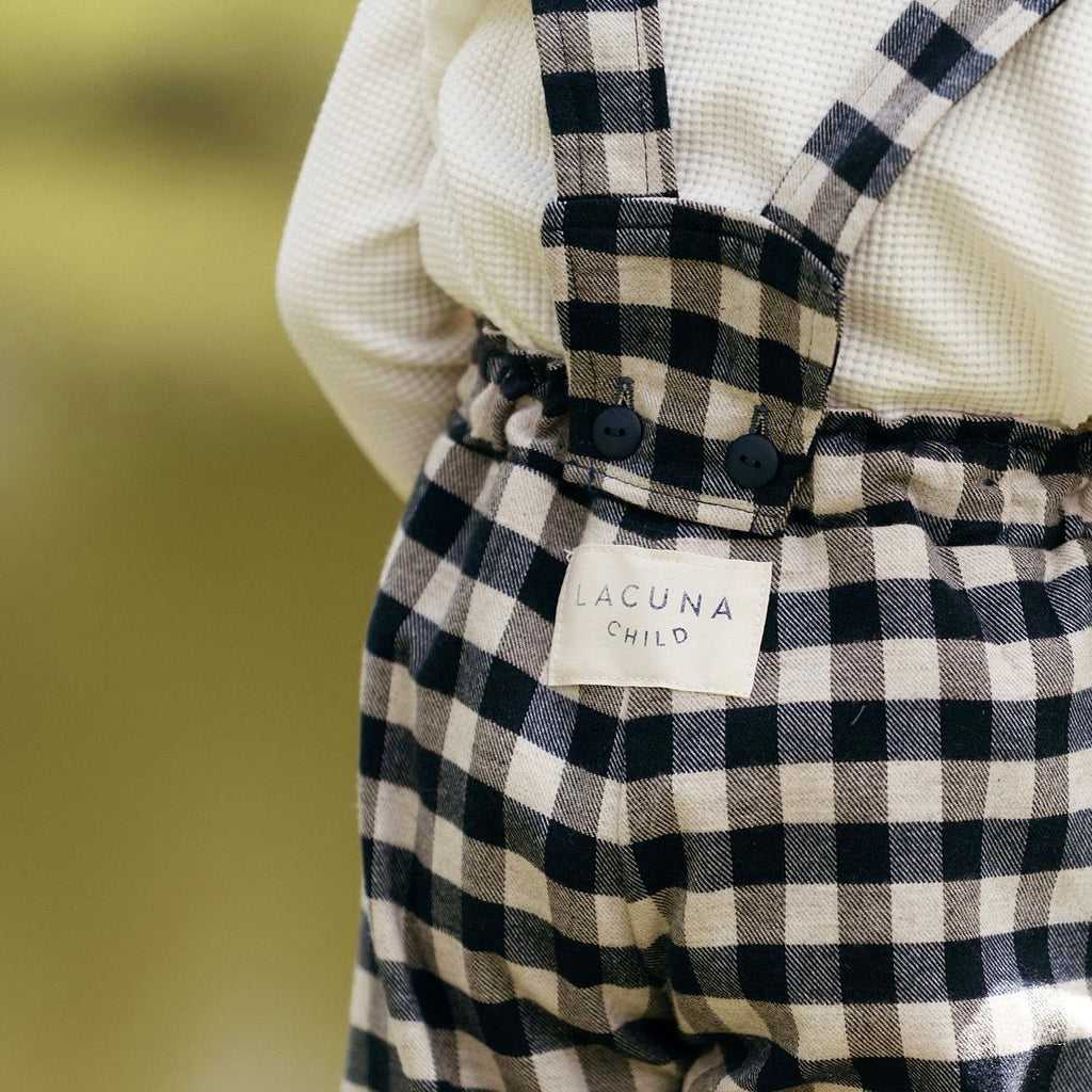 The rear view of the blue brushed cotton and wool gingham check unisex childrens trousers showing the detachable braces and Lacuna Chilld brand label.  Worn with the unisex kids organic jersey offwhite long sleeve top