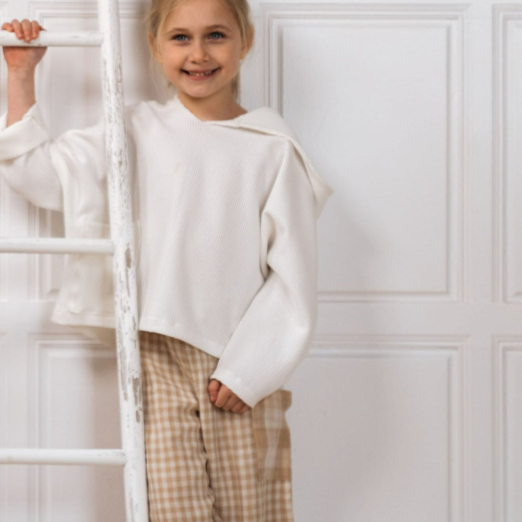 Girl leaning against a white wooden ladder wearing the Alex unisex kids hoodies in offwhite.  Worn too are the Quinn kids unisex sand gingham culottes showing the oversize pocket in the large gingham print on the side of the left leg