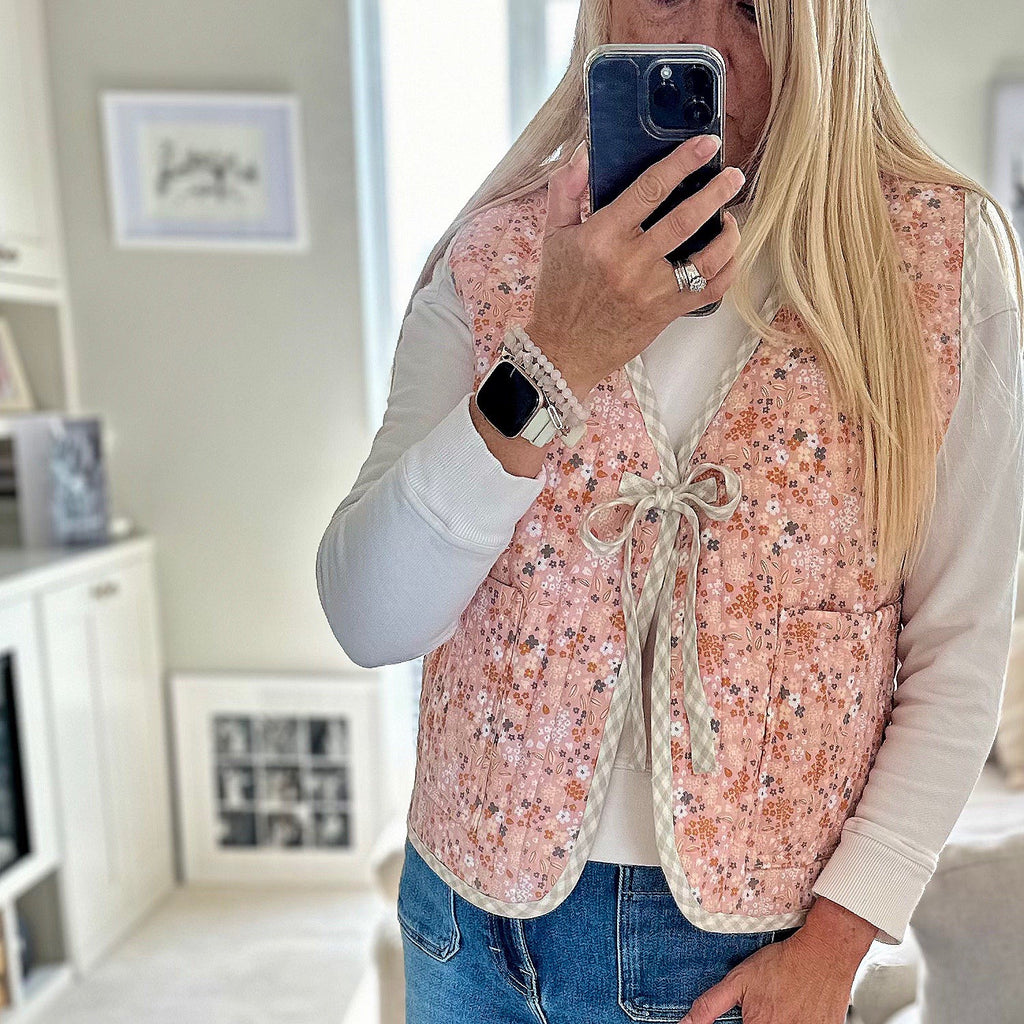 A sleeveless gilet made from quilted cotton.  The base colour for the fabric is peach/pink decorated with a floral pattern of grey, caramel, yellow and offwhite flower clusters.  Reverse of jacket is a pale pink shade with white floral pattern.  Gilet is edged all the way round the jacket with beige gingham linen which is used to provide a bow fastening. 2 oversized square pockets on the front of the gilet can be accessed from the top or the sides of the pockets