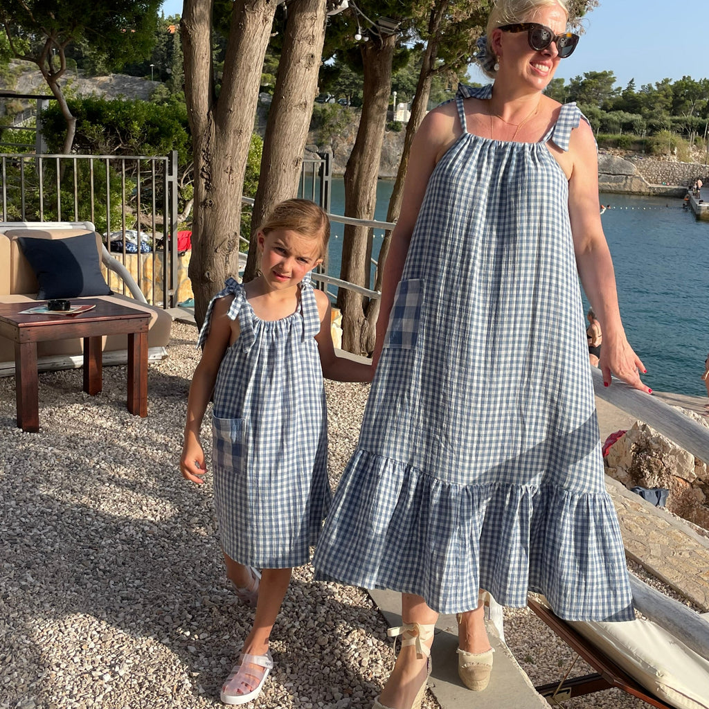 Image of lady and girl both wearing matching blue gingham sundresses with bow tie straps. Ladies dress has gathered tier running round the bottom of the dress, Both dresses have oversized pockets