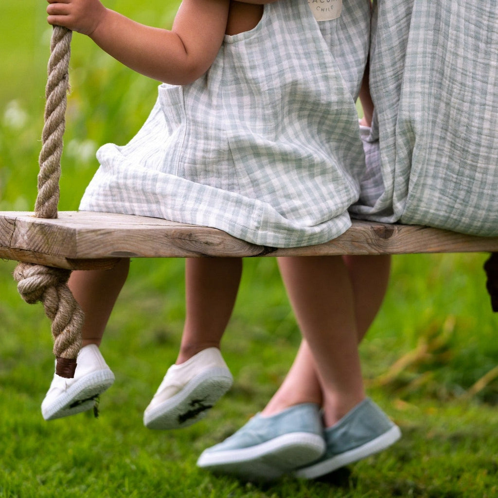 Two girls wearing bow tie summer dress in mint gingham muslin. Showing them both on a swing holding on to the ropes. The perfect child's summer dresses