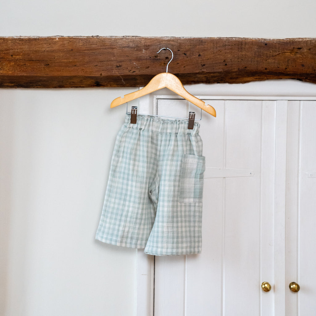 Image shows the Quinn mint gingham infant culottes hanging from a wooden hangar from a wooden beam against a white wooden door