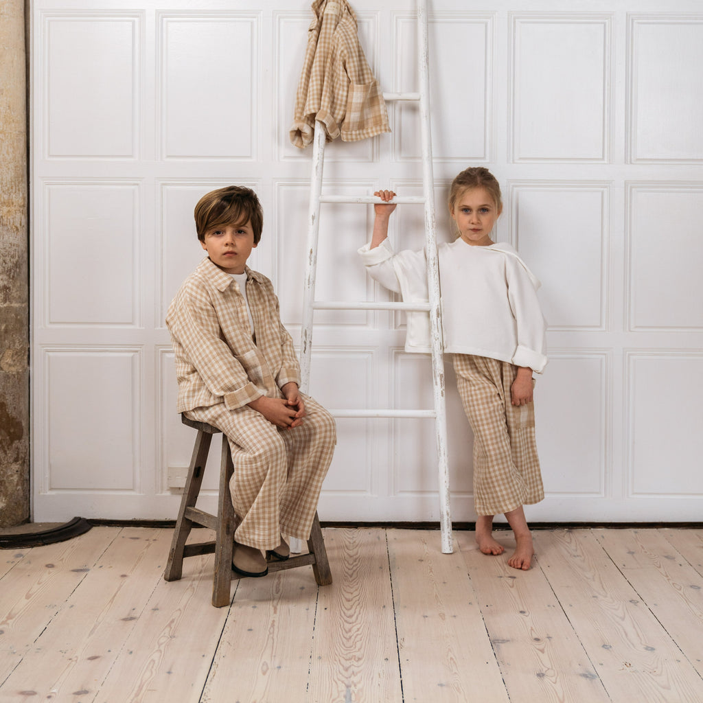 Shown in image are a boy and a girl both wearing the Quinn gingham check culottes in sand.  One child wearing the offwhite Alex hoodie and other child wearing the Bailey gingham check shirt in sand with white tee shirt undernearth.  Hanging from white wooden ladder is the Bailey unisex kids shirt in sand shade 