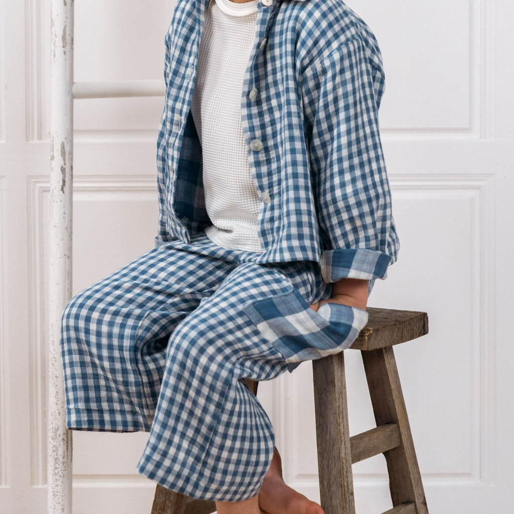 Child sitting on a wooden stool wearing the Bailey blue kids unisex gingham shirt and the Buddy childrens unisex offwhite jersey long sleeve top in offwhite.  Hand in oversized blue gingham large check stitched to the side of the Quinn childrens unisex blue gingham culottes