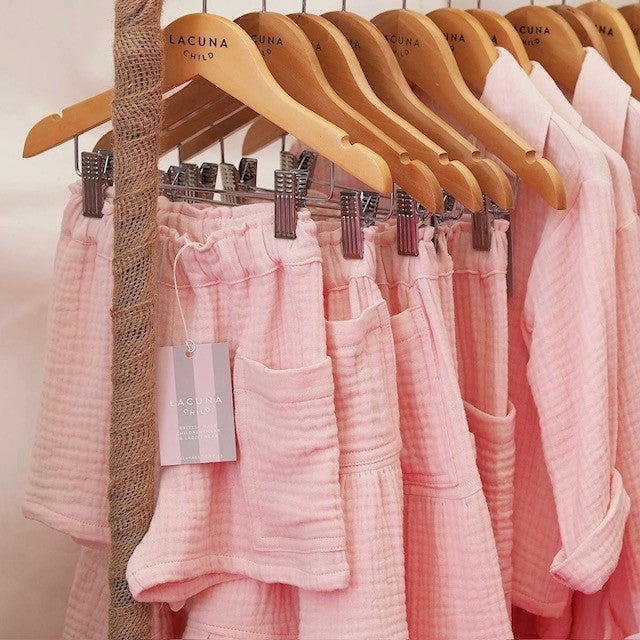 A rail of childrens clothes in a pale pink muslin fabric.  At the front of the rail are the Merin shorts with adjustable elasticated waist and an oversized pocket.  The Pink and Grey stripe Lacuna Child swing tag is also displayed