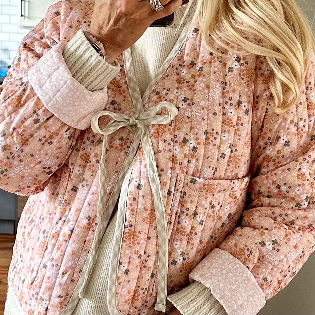 Ladies cotton quilted floral jacket.  Base colour of jacket is peach/pink with floral pattern.  Inside pattern is pale pink with white floral pattern.  Oversized roll up sleeves showing reverse pink fabric.  Jacket is edged with beige linen check which is used for fastening ties.  2 oversized pockets on the front which allow hands to access from the side or from the top of the jacket
