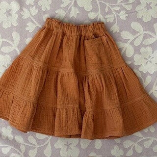 Girls knee length cotton muslin tiered skirt in soft orange.  Shown with a single oversized pocket