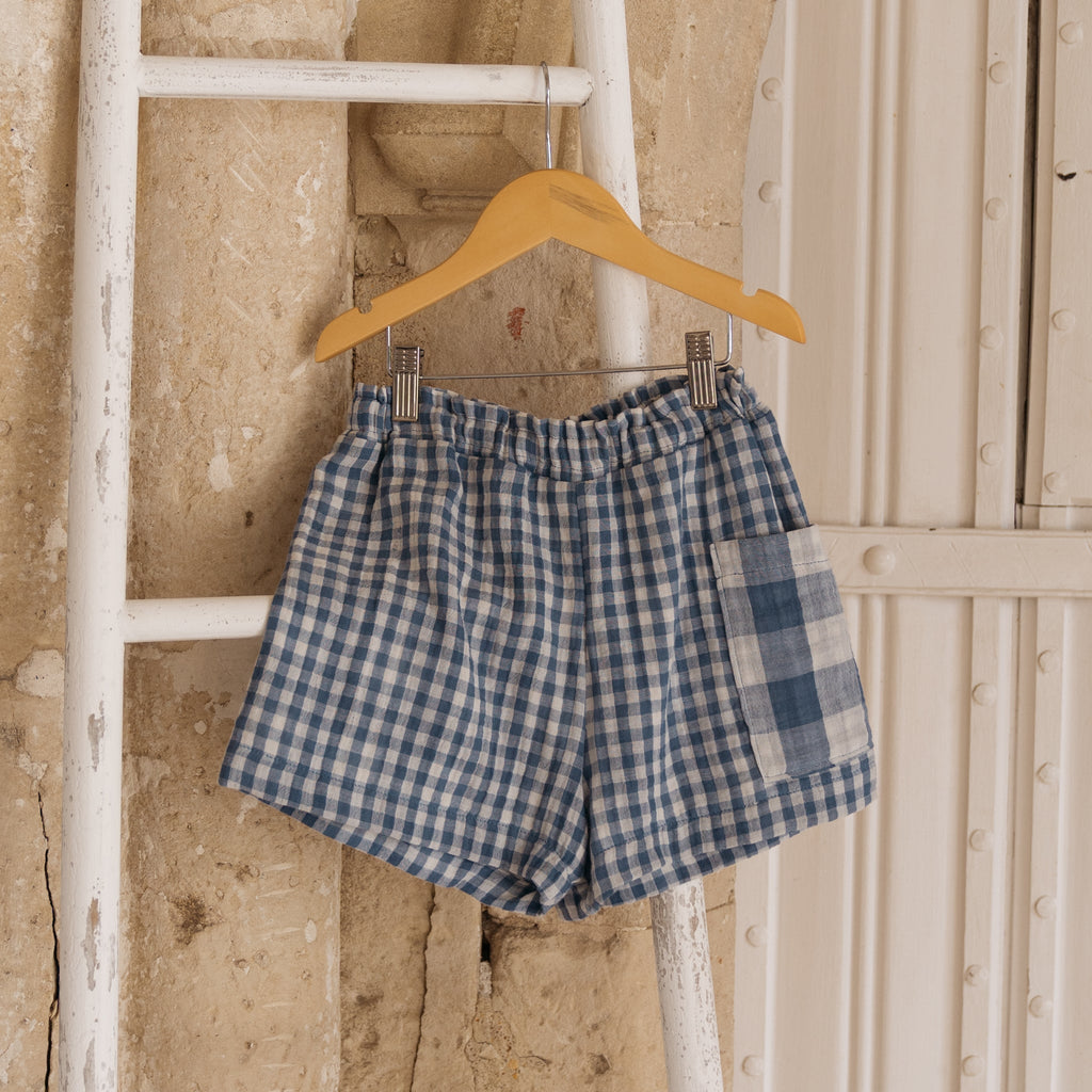 The Merin kids unisex shorts in blue gingham with adjustable elasticated waist with oversized pocket in large blue gingham check clipped to a wooden hangar 